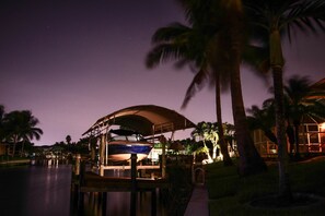 A boat docked under a covered area with palm trees and a building in the background. - Dream villa in top location on the wide canal & large pool area with jacuzzi (Cape Coral)