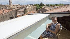 Een vrouw zit op een balkon met uitzicht op een stadsgezicht, inclusief een toren en gebouwen met terracotta daken.
