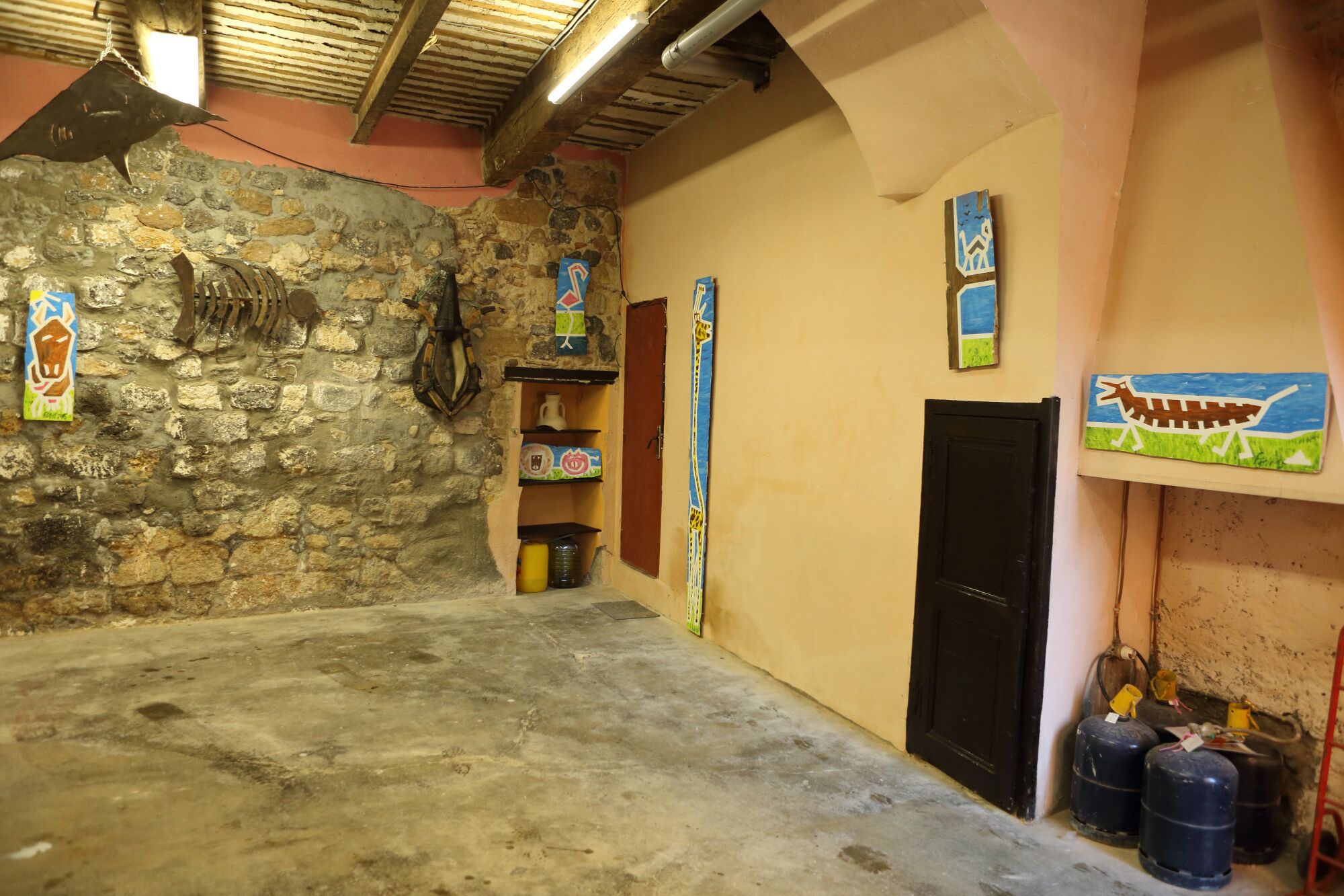 Ein Zimmer mit einer Steinwand, einer schwarzen Tür und einem Betonboden. Bunte Gemälde schmücken die Wand und ein Regal mit Gegenständen ist vorhanden.