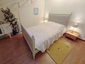 A bedroom with a white bed, a bedside table with a lamp, a wooden nightstand, a small potted plant, and a wall-mounted picture.