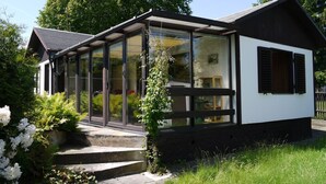 Una casa moderna con un invernadero de vidrio, una terraza de madera y un jardín con flores blancas.