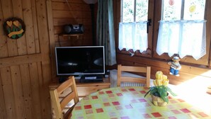 L’intérieur d’un chalet en bois, avec un téléviseur posé sur un meuble, une table recouverte d’une nappe colorée et une figurine d’ours en peluche sur le rebord de la fenêtre.