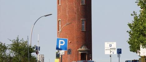 A brick tower with a dome and a balcony, surrounded by a paved area with cars and street signs.