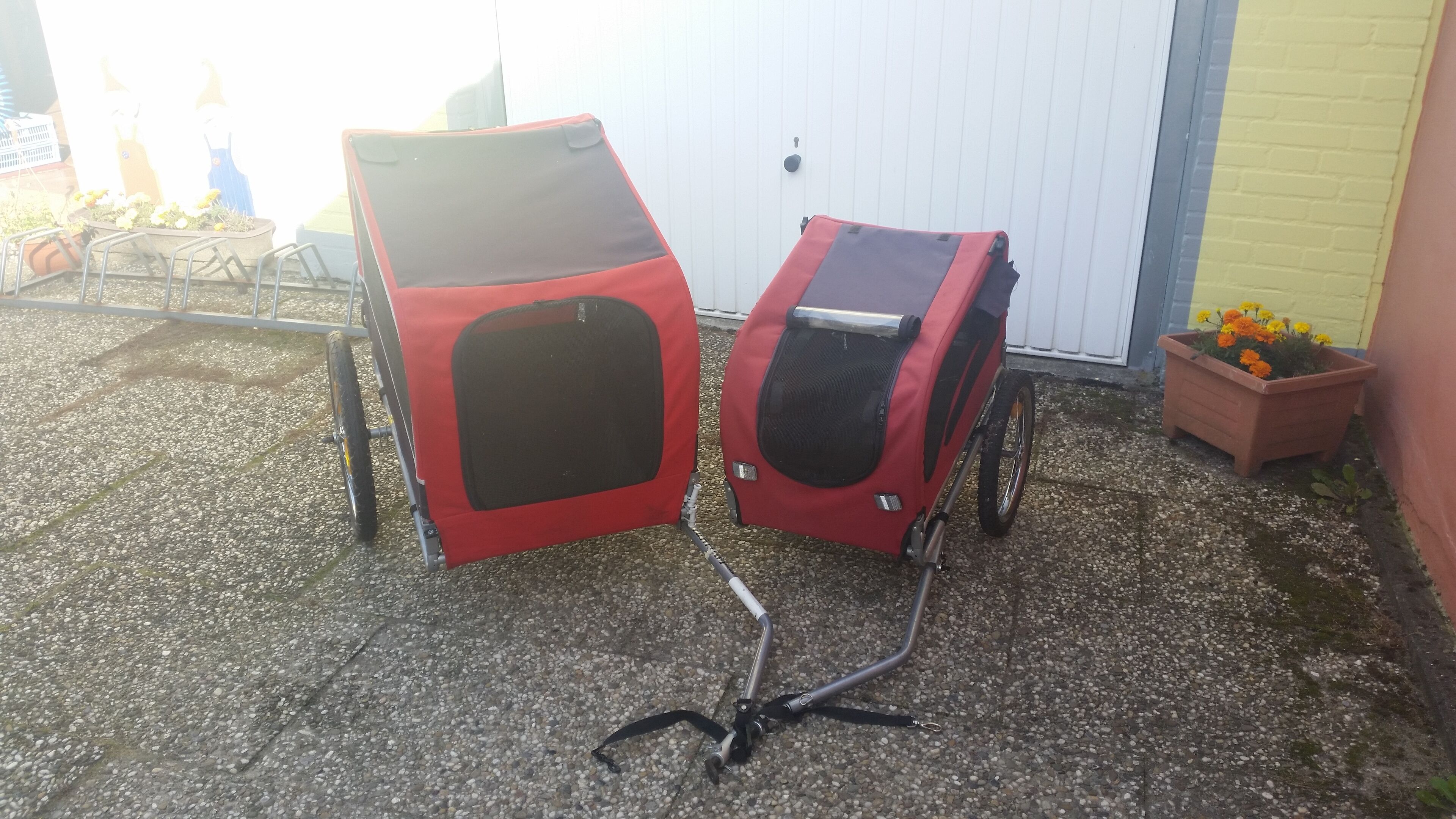 Two red baby strollers with black seats and a metal frame, parked on gravel.