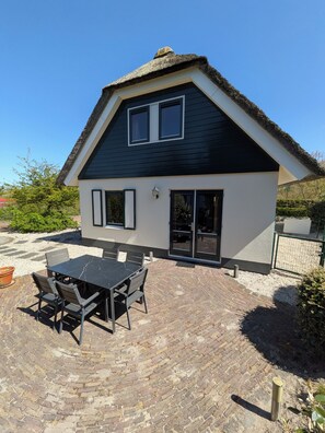 Une maison avec un toit de chaume, une terrasse avec une table et des chaises, et un chemin en briques.