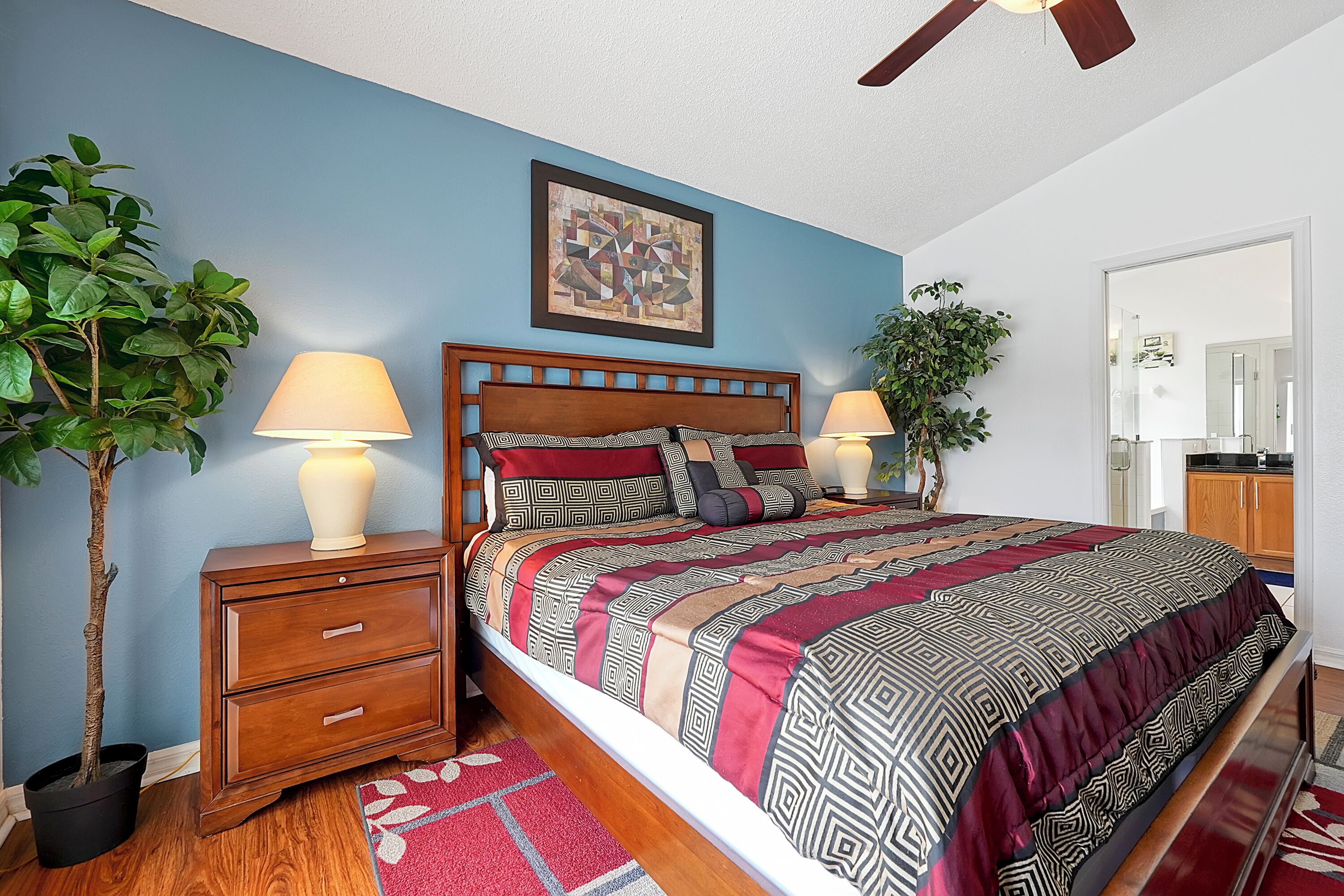 A bedroom with a wooden bed, two bedside tables with lamps, a large potted plant, and a painting on the wall.