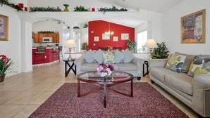 Una sala de estar con una pared de acento roja, una mesa de centro de vidrio con flores, un sofá gris y una cocina al fondo.