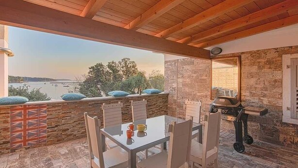 Eine überdachte Terrasse im Freien mit Tisch, Stühlen, einem Grill und Blick auf ein Gewässer.
