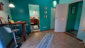 A teal-walled room with a kitchen area, a dining table with chairs, and a patterned rug.
