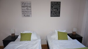 Ein Hotelzimmer mit zwei Betten, jeweils mit weißer Bettwäsche und grünen Kissen. Zwei Nachttische mit Lampen, und motivierende Bilder an der Wand.