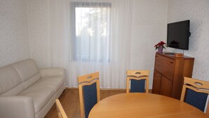 Ein Zimmer mit einem beigefarbenen Sofa, einem Holzesstisch mit vier Stühlen, einem Fernseher an der Wand und einem Fenster mit durchsichtigen Vorhängen.