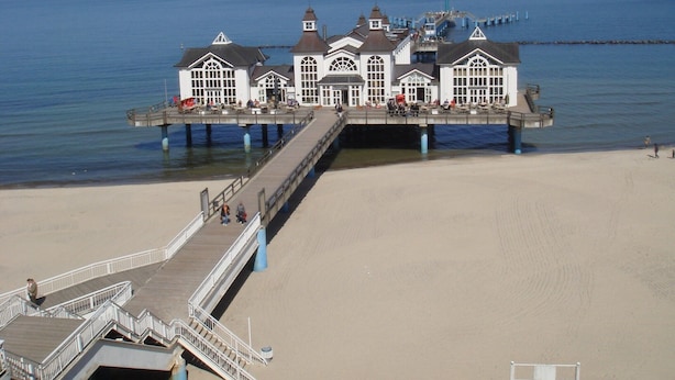 A pier extending into the sea with a white building at the end, featuring multiple windows and a staircase leading to the beach.
