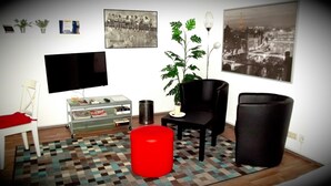 Un salon comprenant un téléviseur à écran plat, une table basse en verre, un pouf rouge, un fauteuil noir, une plante en pot et un tapis à motifs.
