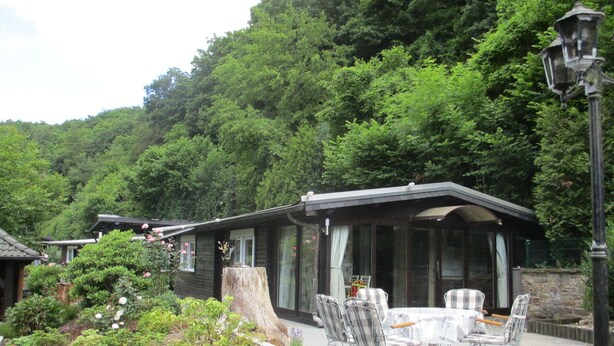 Ein überdachter Sitzbereich im Freien mit Tisch und Stühlen, umgeben von üppiger Vegetation und einem bewaldeten Hang.