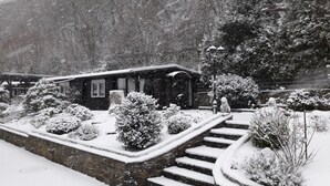 Ein mit Schnee bedeckter Garten mit einer Steinmauer, Stufen zu einem Holzhaus und schneegefüllten Bäumen.