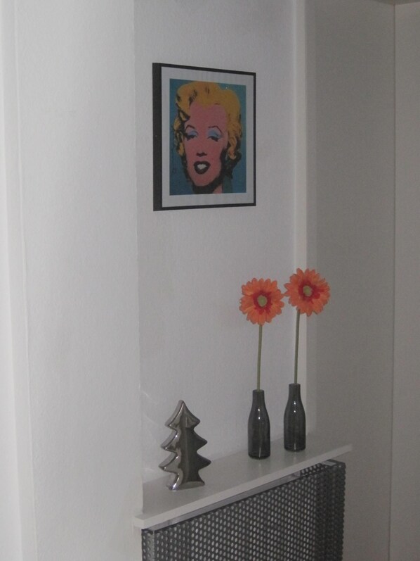 Um retrato pop art emoldurado numa parede branca, uma árvore de metal decorativa e dois vasos com flores laranjas numa prateleira branca.