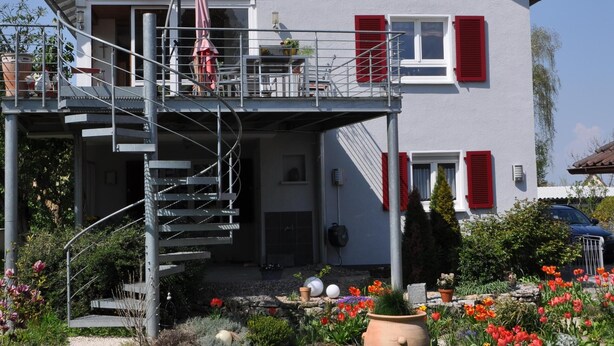 Een twee verdiepingen tellend huis met een balkon, een spiraaltrap en een tuin met bloemen.