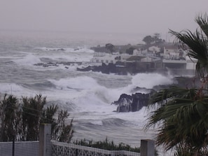 Vista costera con olas del mar rompiendo contra una costa rocosa, un edificio blanco con múltiples ventanas y una palmera en primer plano.