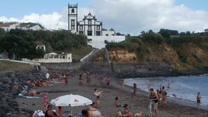 Une plage où des gens se prélassent au soleil et se baignent, un bâtiment blanc avec un clocher en arrière-plan, et une côte rocheuse.