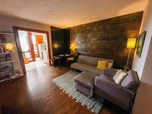 A living room with a sofa, a coffee table, and a bookshelf. There is a dining area with a table and chairs.