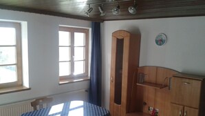 A room with a round table covered in a checkered tablecloth, a tall wooden cabinet, a window with a blue curtain, and a wall clock.