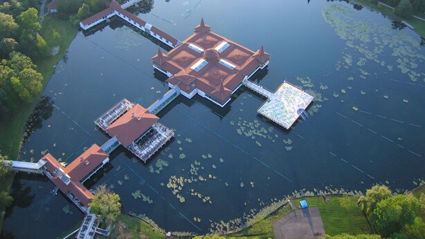 Une vue aérienne d’une vaste étendue d’eau circulaire, avec une île centrale sur laquelle se trouve un bâtiment traditionnel doté d’un toit rouge et de plusieurs annexes.