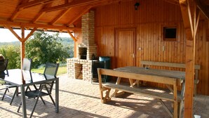 Un espace extérieur abrité, doté d’une table et de chaises en bois, d’une cheminée en pierre et offrant une vue sur le paysage environnant.