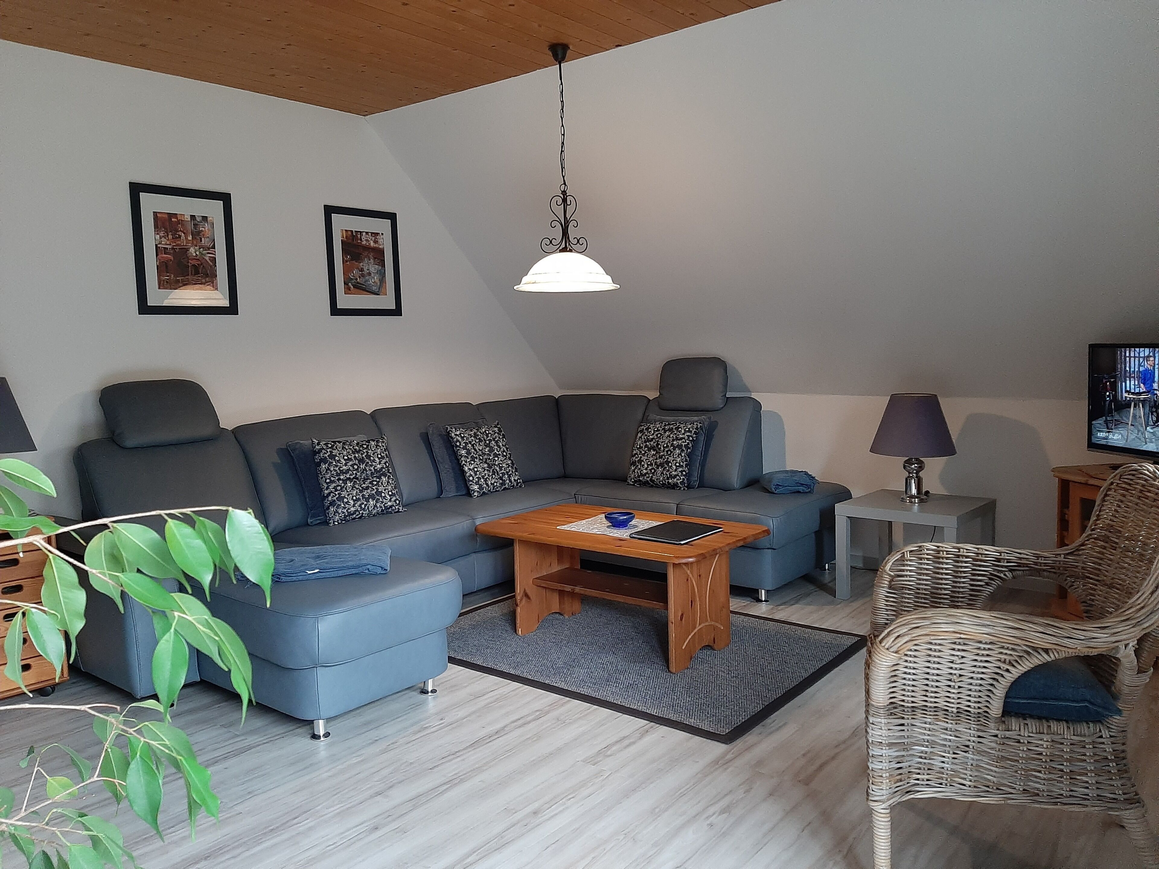 Ein modernes Wohnzimmer mit einem grauen Ecksofa, einem Holztisch, einem Korbsessel und einem Fernseher auf einem Lowboard.