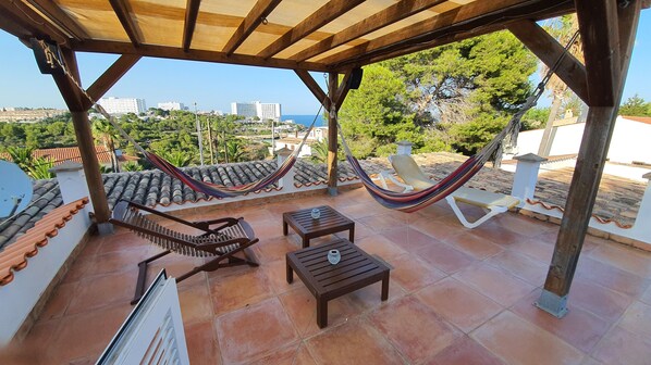 A covered patio with a hammock, wooden lounge chairs, and a tiled floor.