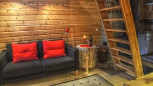 A cozy log cabin interior with a wooden sofa, red cushions, a small wooden table with a candle, and a ladder leading to a kitchen area. - Quaint holiday home in the forests of Lapland by the river (Sorsele)