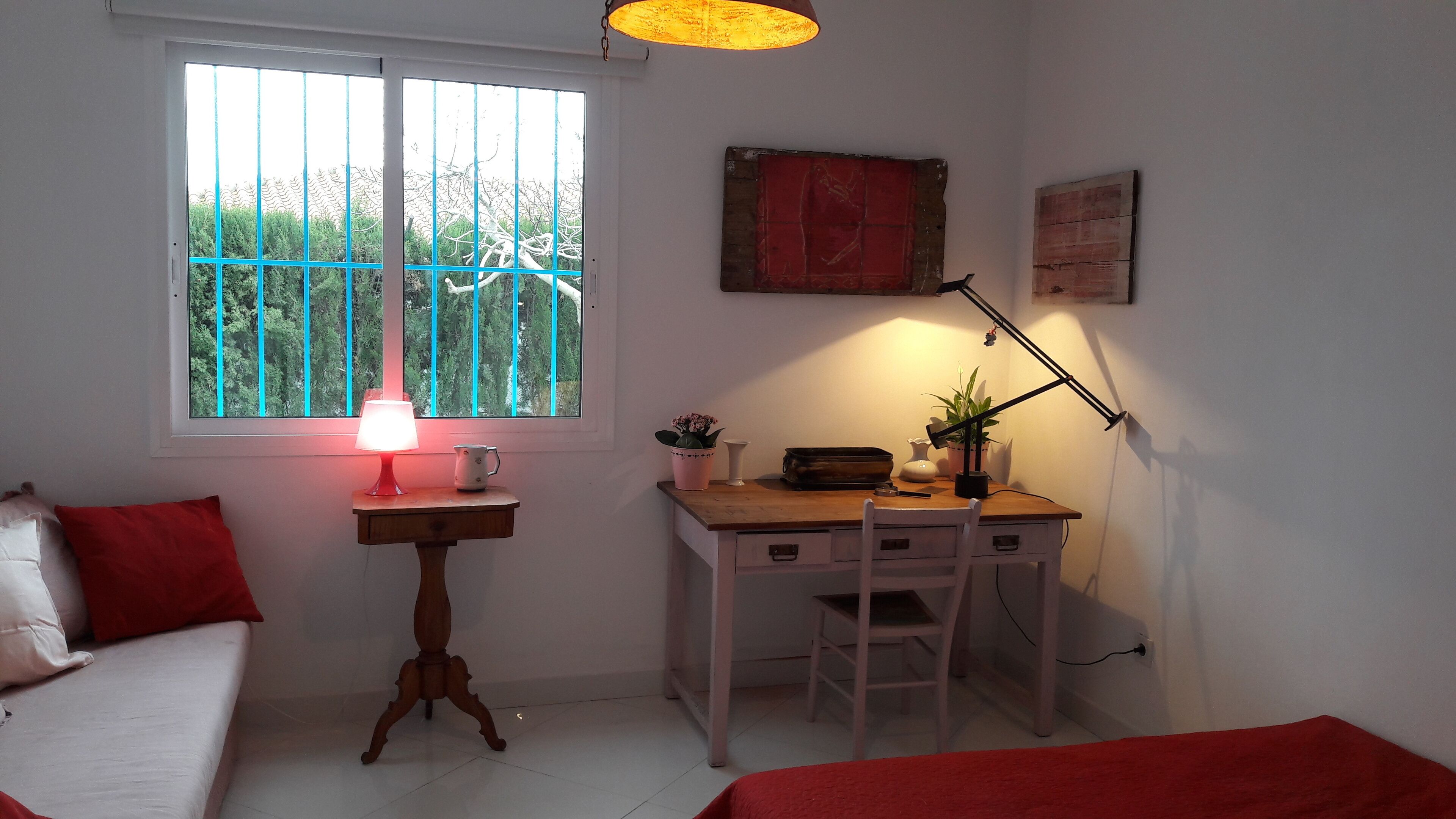 Un dormitorio con cama, escritorio con lámpara, ventana con persianas y una pequeña mesa con planta.