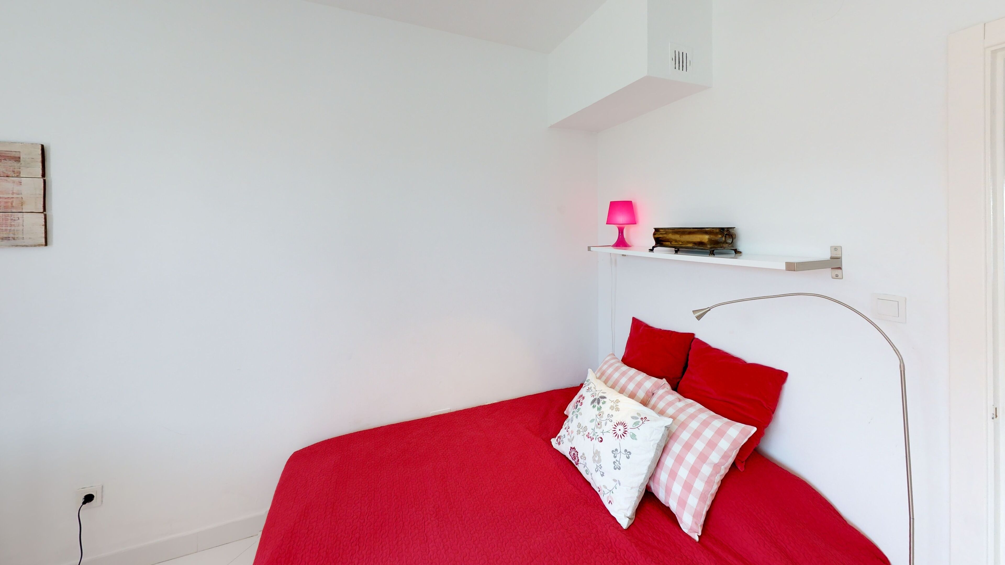 A bedroom with a red bedspread, a white wall, and a pink lamp on a shelf.