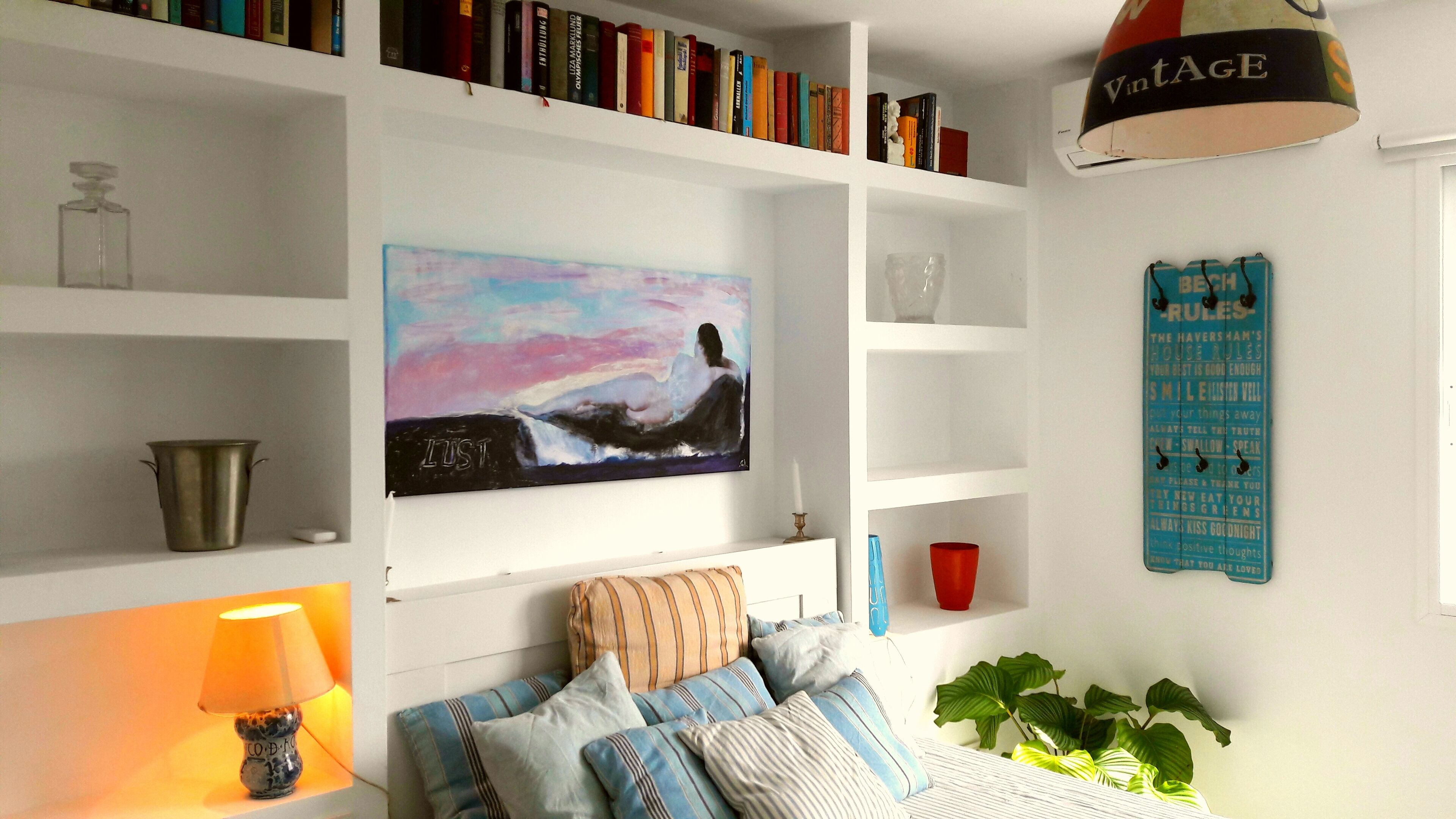 A bedroom with a bed, pillows, a bedside table with a lamp, a painting, shelves with books, a vase, and a wall-mounted sign.