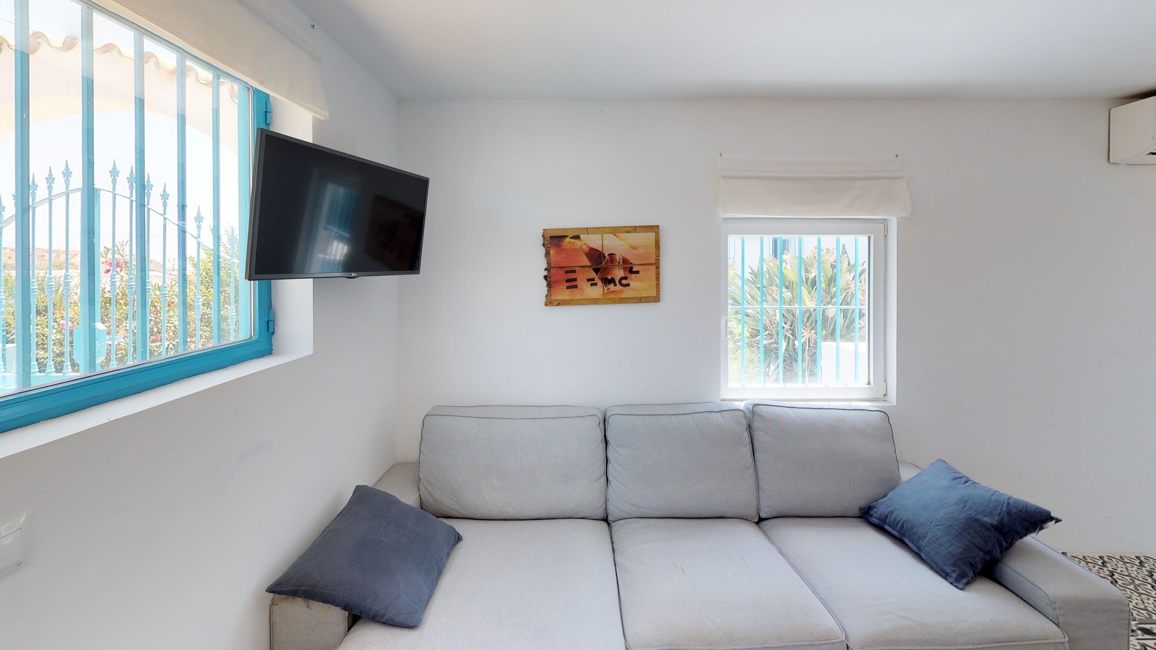 Ein Wohnzimmer mit einer Couch, einem an der Wand montierten Flachbildfernseher und einem Fenster mit Jalousien.