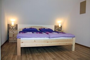 Ein Einzelbett mit Holzrahmen und Kopfteil, flankiert von zwei Nachttischen mit Lampen. Der Raum hat Holzboden und schlichte Wände.