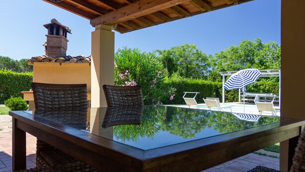 Une table à plateau en verre, des chaises en osier, une cheminée en briques et un parasol au bord de la piscine, le tout dans un cadre extérieur ensoleillé.