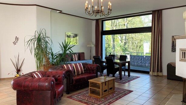 A living room with a sofa, a dining table, chairs, a chandelier, a large window with curtains, and a view of trees. - Villa Warzenburg, apartment 2 with panoramic view, balcony and fireplace. Vacation with a dog. (Friedrichroda)