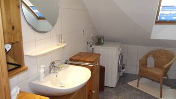 Une petite salle de bain avec un meuble-lavabo en bois, un lavabo blanc, un miroir, un lave-linge et une chaise en osier.
