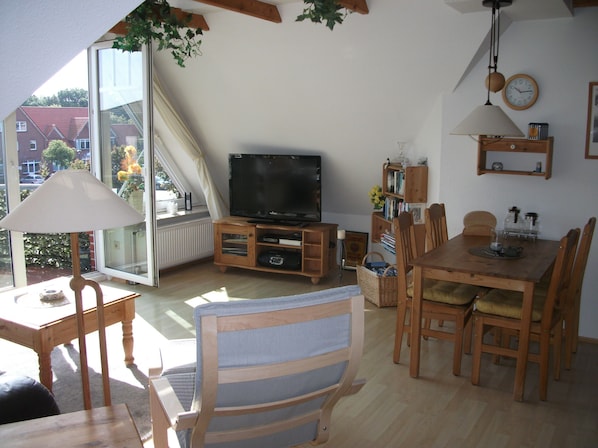A cozy living room with a television, wooden furniture, a dining table, and a large window offering a view of the outside.
