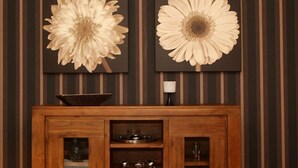 Ein Holzkabinent mit Glastüren und Regalen, ein kleiner Tisch mit einer Schale und einer Lampe, sowie zwei große Blumenbilder an der Wand.