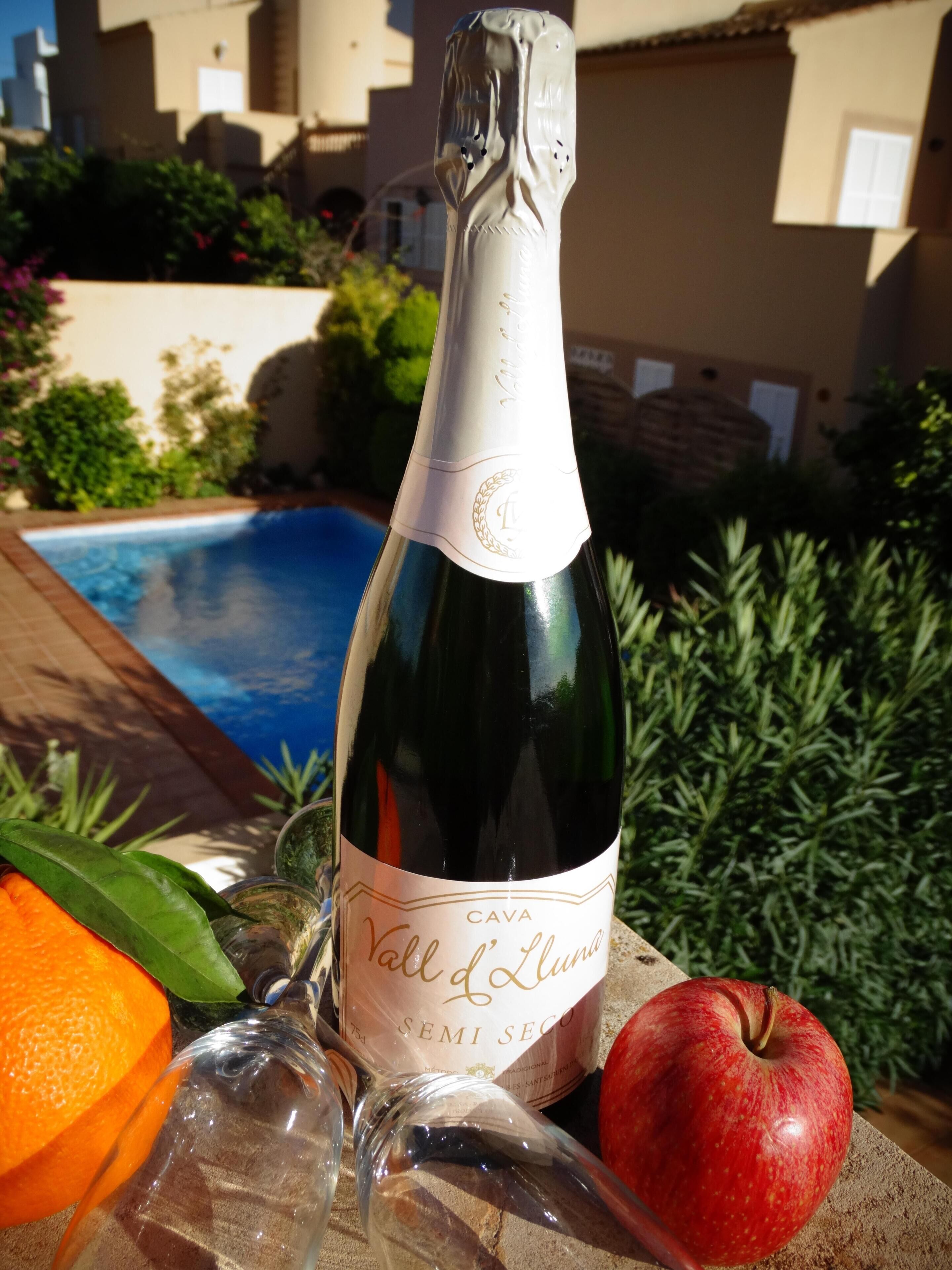 Une bouteille de vin CAVA, un verre, une pomme rouge et une orange coupée en deux, posés sur une table près d’une piscine.