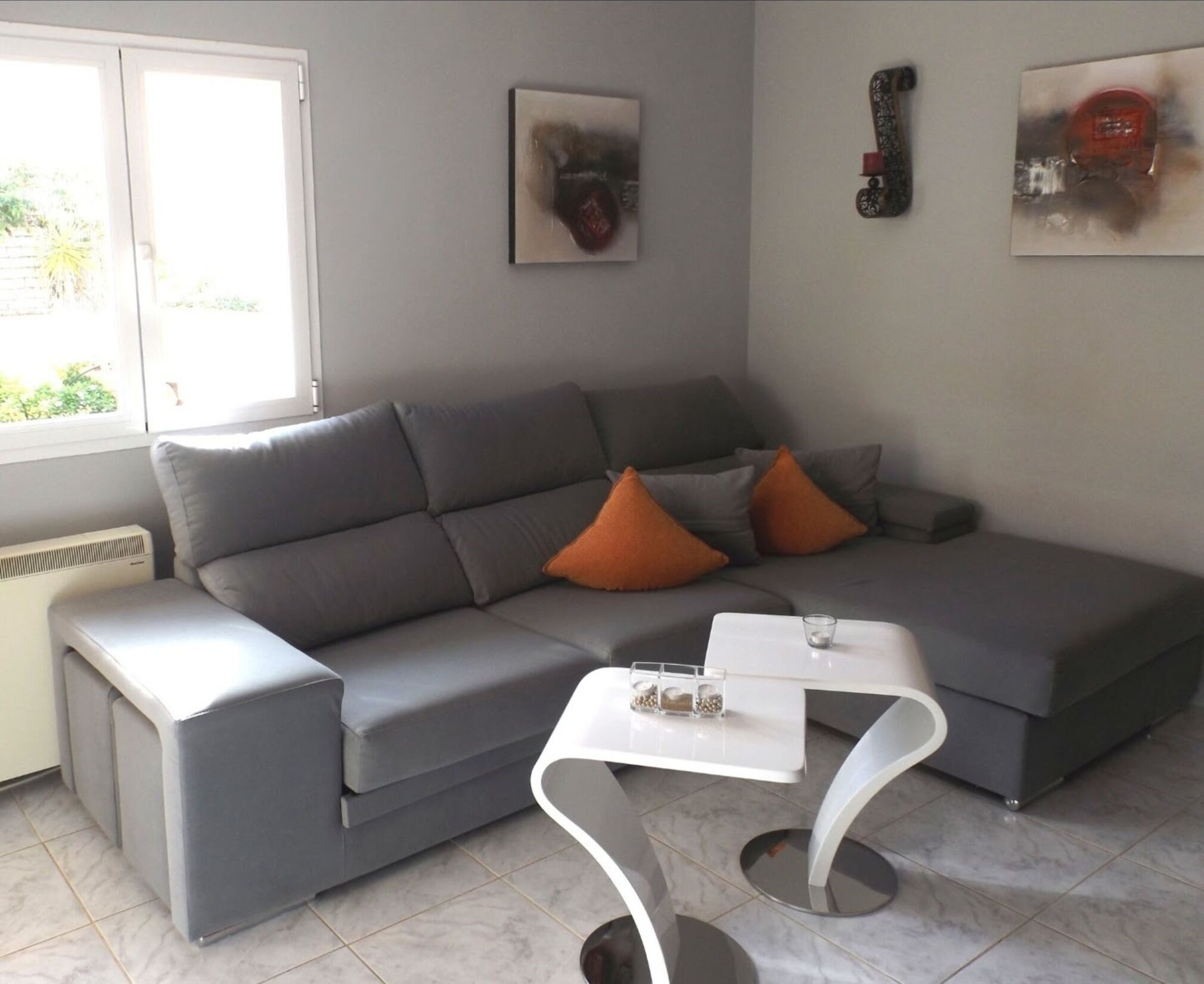 Een moderne woonkamer met een grijze bank, oranje kussens, een witte salontafel en abstracte kunstwerken aan de muren.