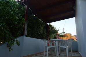 An outdoor patio with a round table and white plastic chairs, surrounded by a low white wall and dense green foliage.