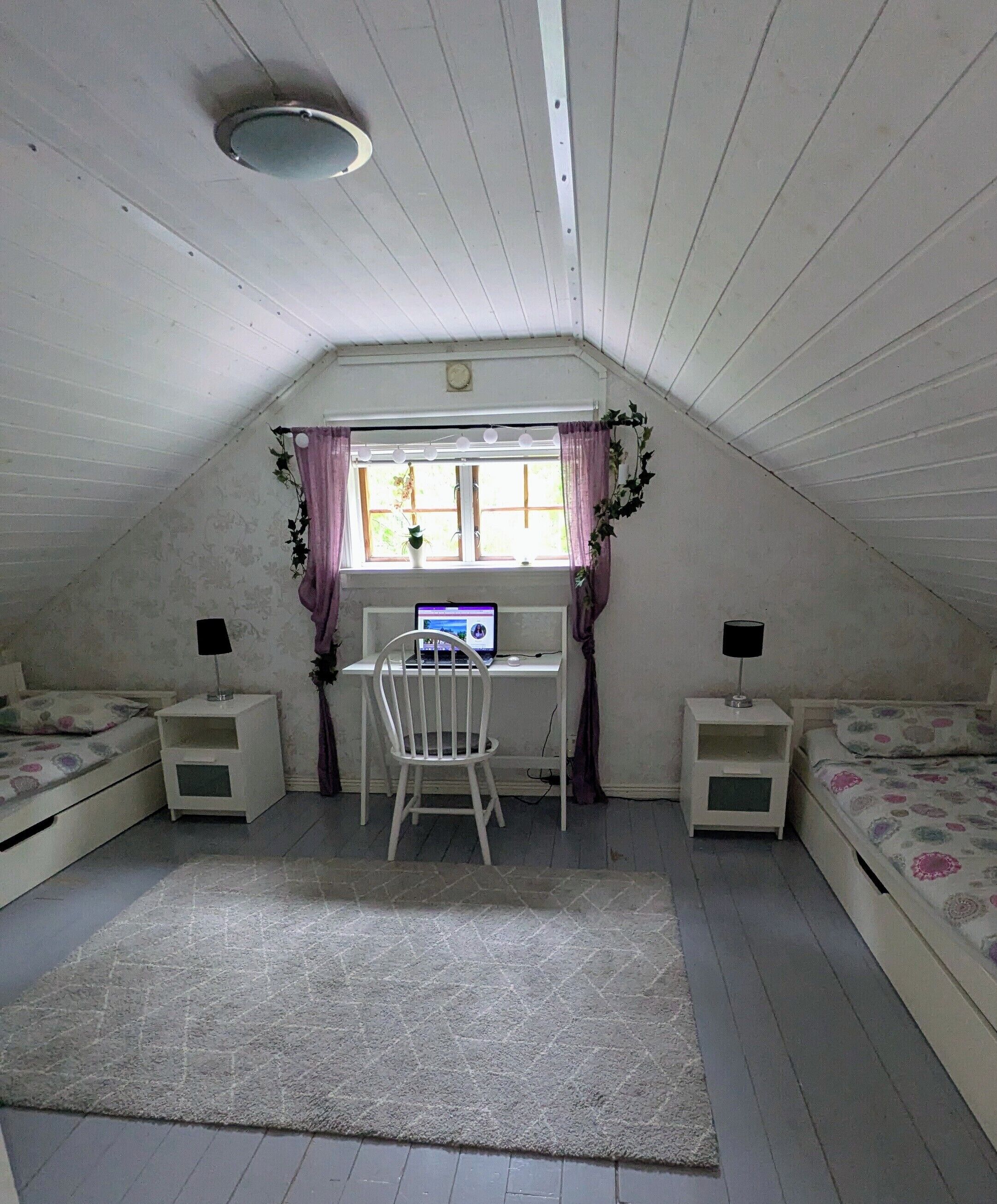 Een kamer met schuine plafonds, een bureau met computer, twee nachtkastjes, een stoel en een bed met bloemenprint beddengoed.