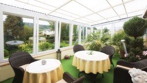 A conservatory with round tables and wicker chairs, large windows, and a view of a garden. - Holiday Apartment "Pharmacy" with Weinbergblick (Trittenheim)