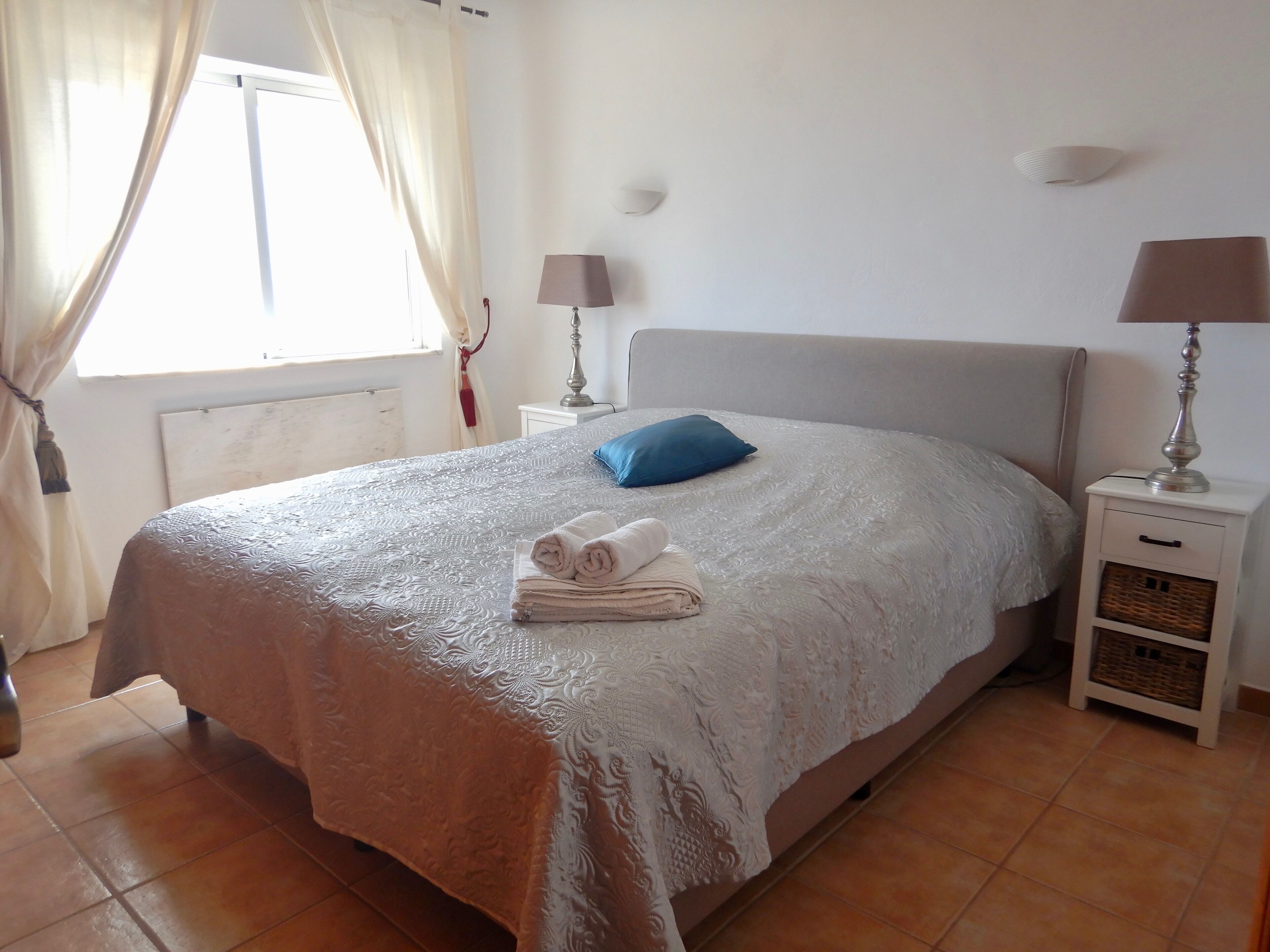 A neatly made bed with a white bedspread, two pillows, and folded towels on the side. There are two bedside tables with lamps, a window with curtains, and a radiator on the wall.