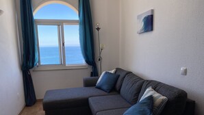 Un salon moderne avec un canapé d’angle gris, une petite table et une vue sur la mer, visible à travers une fenêtre ornée de rideaux turquoise.