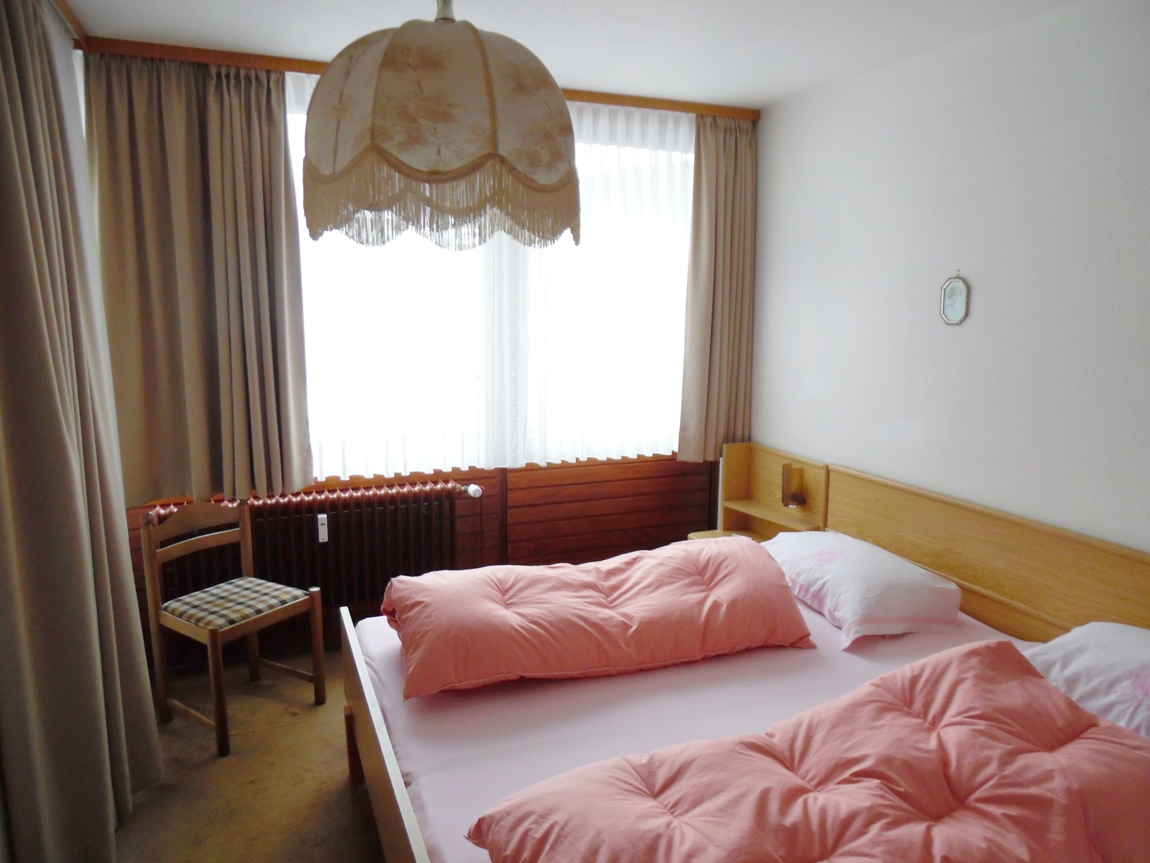 A hotel room with two beds, a wooden headboard, a chair, a radiator, and a window with curtains.