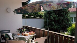 Un espace de restauration en plein air avec une table dressée pour deux personnes, des chaises, un parasol à motifs floraux et une vue sur une clôture en bois et de la verdure.