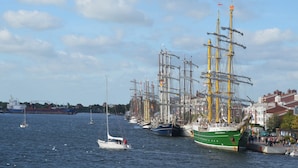 Un port où sont amarrés plusieurs grands voiliers, un petit voilier et des bâtiments qui bordent la promenade.